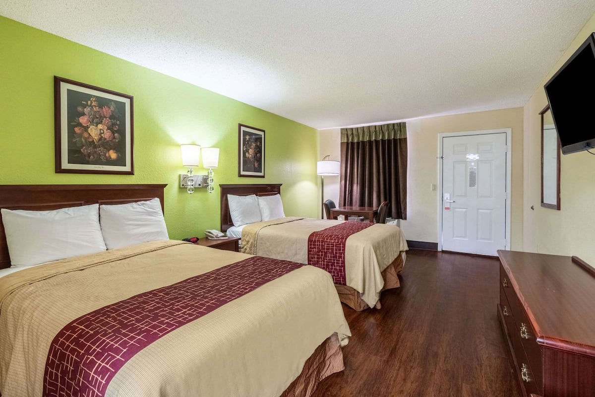 Quarto de hotel com duas camas, uma escrivaninha, uma cadeira, uma televisão e uma porta.
