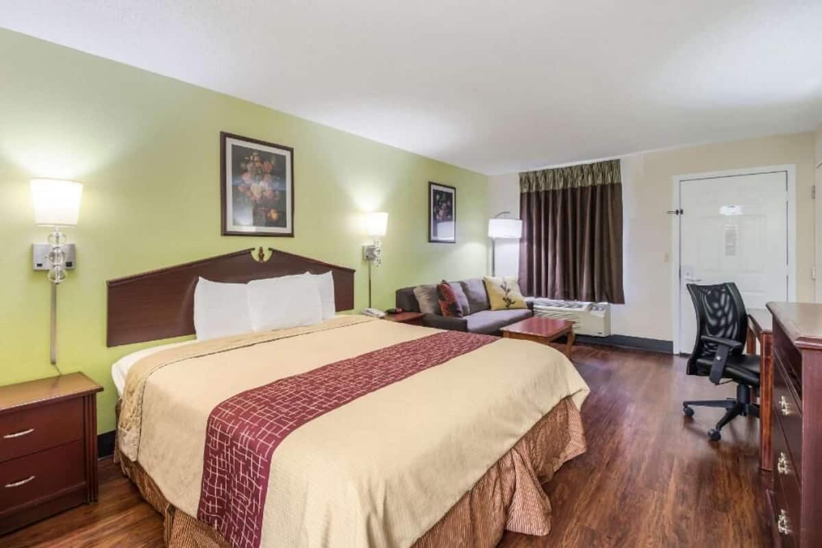 Quarto de hotel com uma cama grande, uma escrivaninha, uma cadeira, um abajur e uma porta.