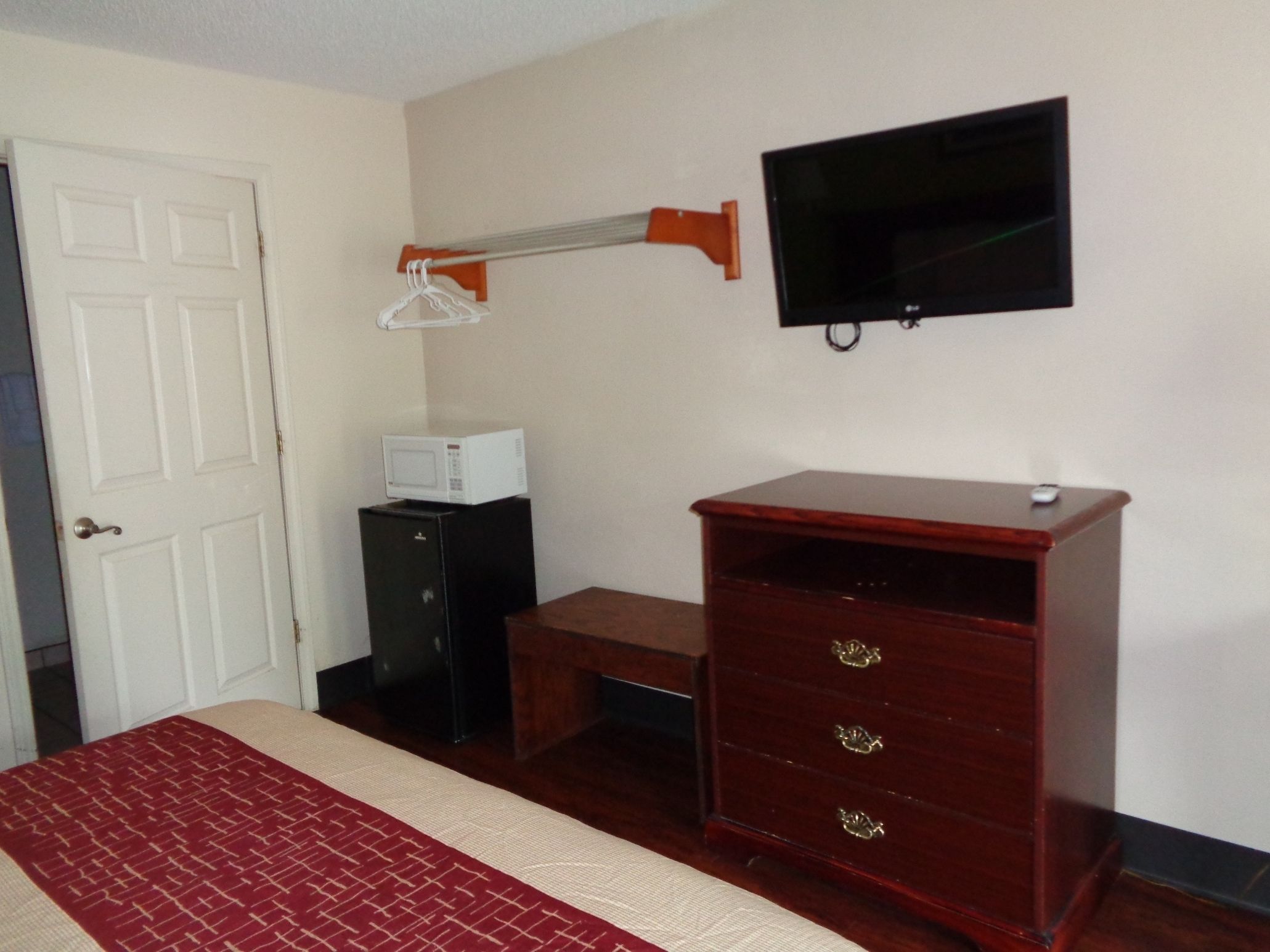 A room with a bed, a wooden dresser, a desk, a microwave, a television, and a door.