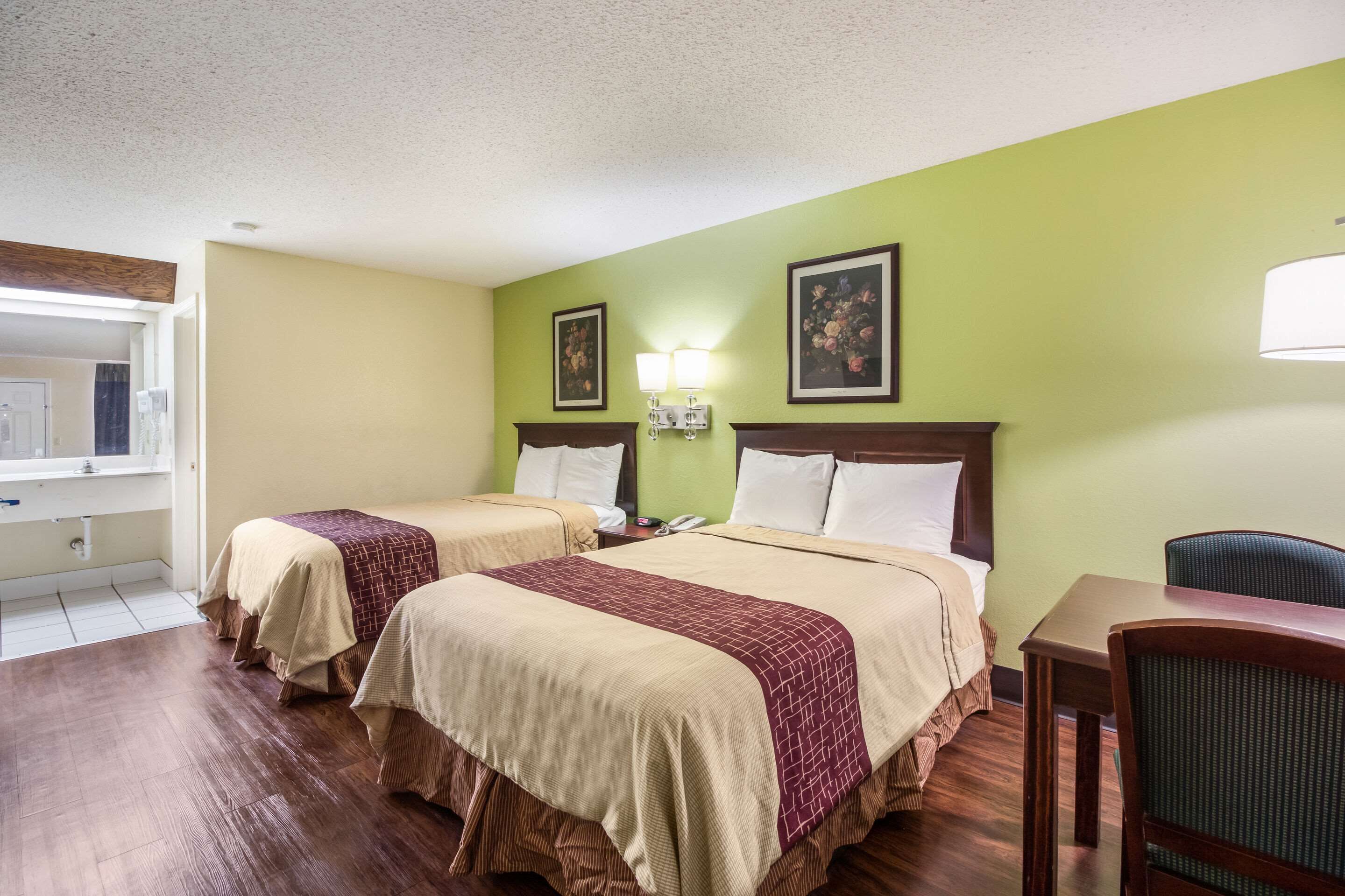 Quarto de hotel com duas camas, uma mesa de madeira, uma cadeira, um criado-mudo e uma luminária na parede.