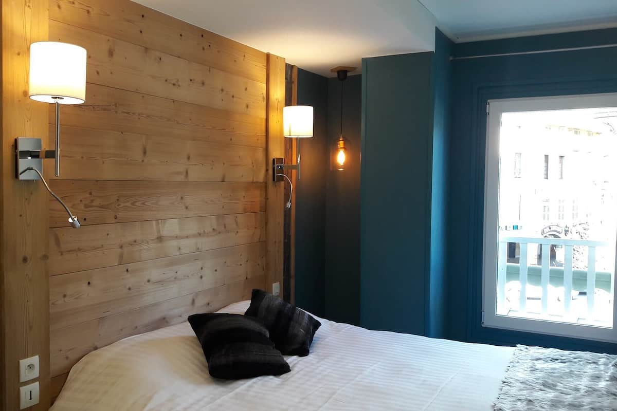 Habitación de hotel con cabecera de madera, dos lámparas de pared, cama con ropa blanca y almohadas negras, y vista a un balcón a través de una ventana.