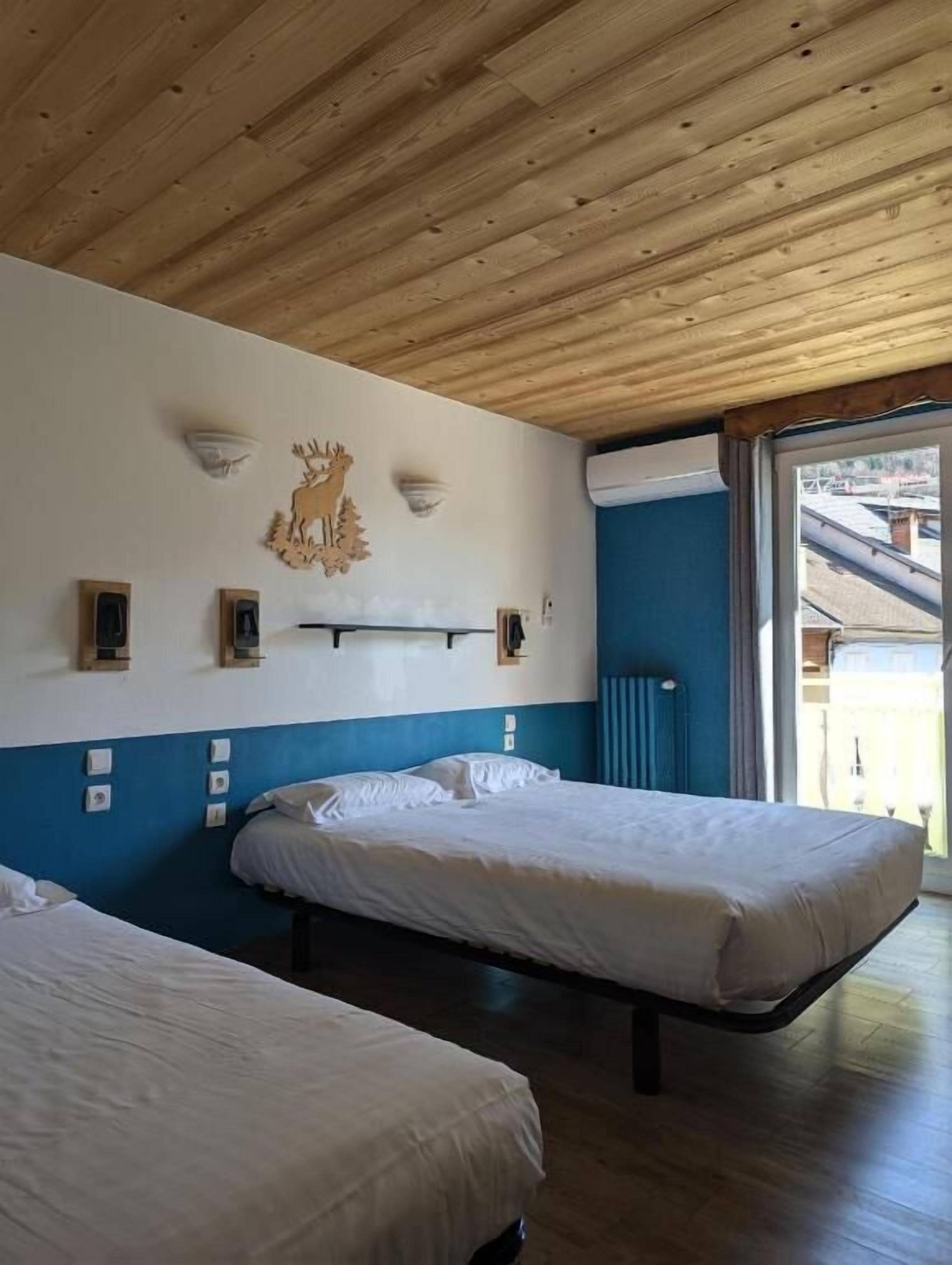 A room with two beds, wooden ceiling, wall-mounted light fixtures, and a window with a view of buildings.