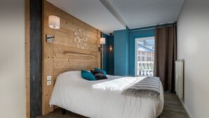 A modern hotel room with a wooden headboard, a large bed with white bedding, teal and black pillows, and a view of buildings through a window with curtains. - Hotel du Midi (Thones)