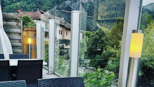 An outdoor dining area with a table and chairs, a menu on the table, and a view of a mountain and greenery in the background. - Hotel du Midi (Thones)