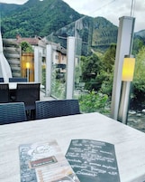 Un área de comedor al aire libre con mesa y sillas, un menú sobre la mesa y vista a una montaña y vegetación de fondo.