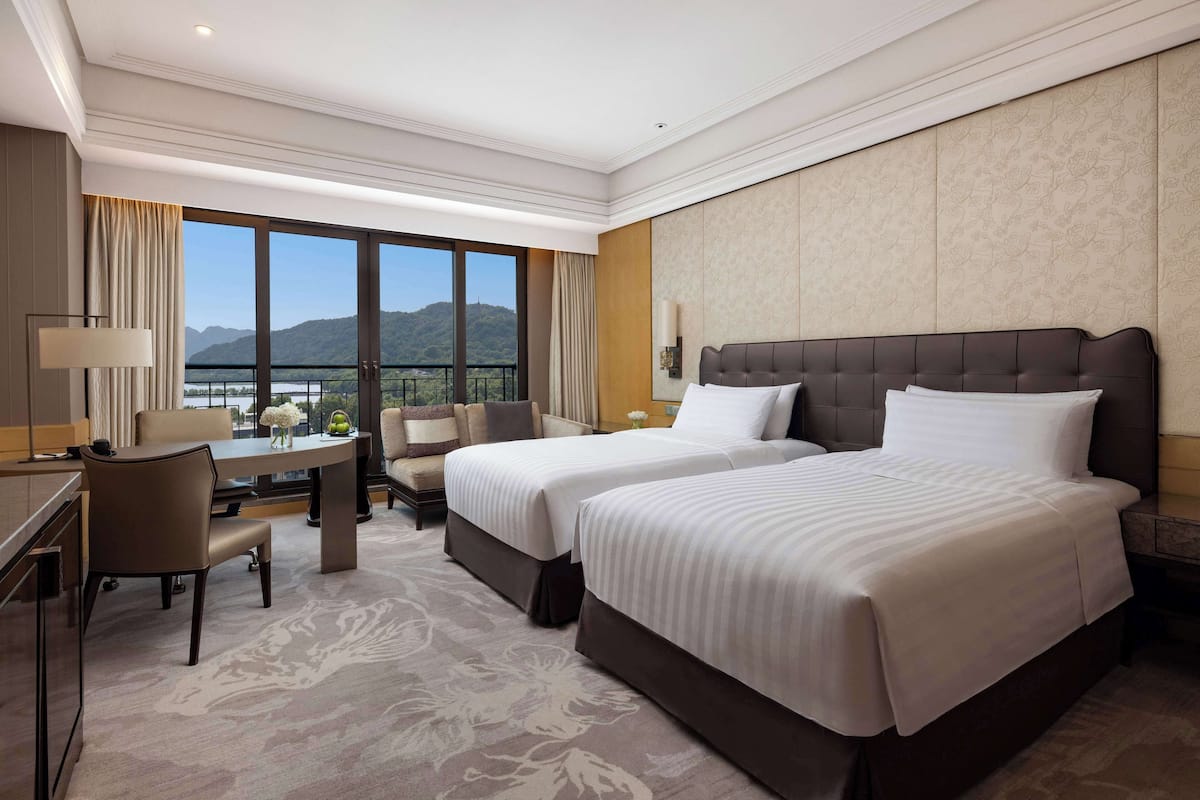 Ein Hotelzimmer mit zwei Betten, einem Schreibtisch und einem Sessel. Ein großes Fenster bietet Blick auf die Berge.