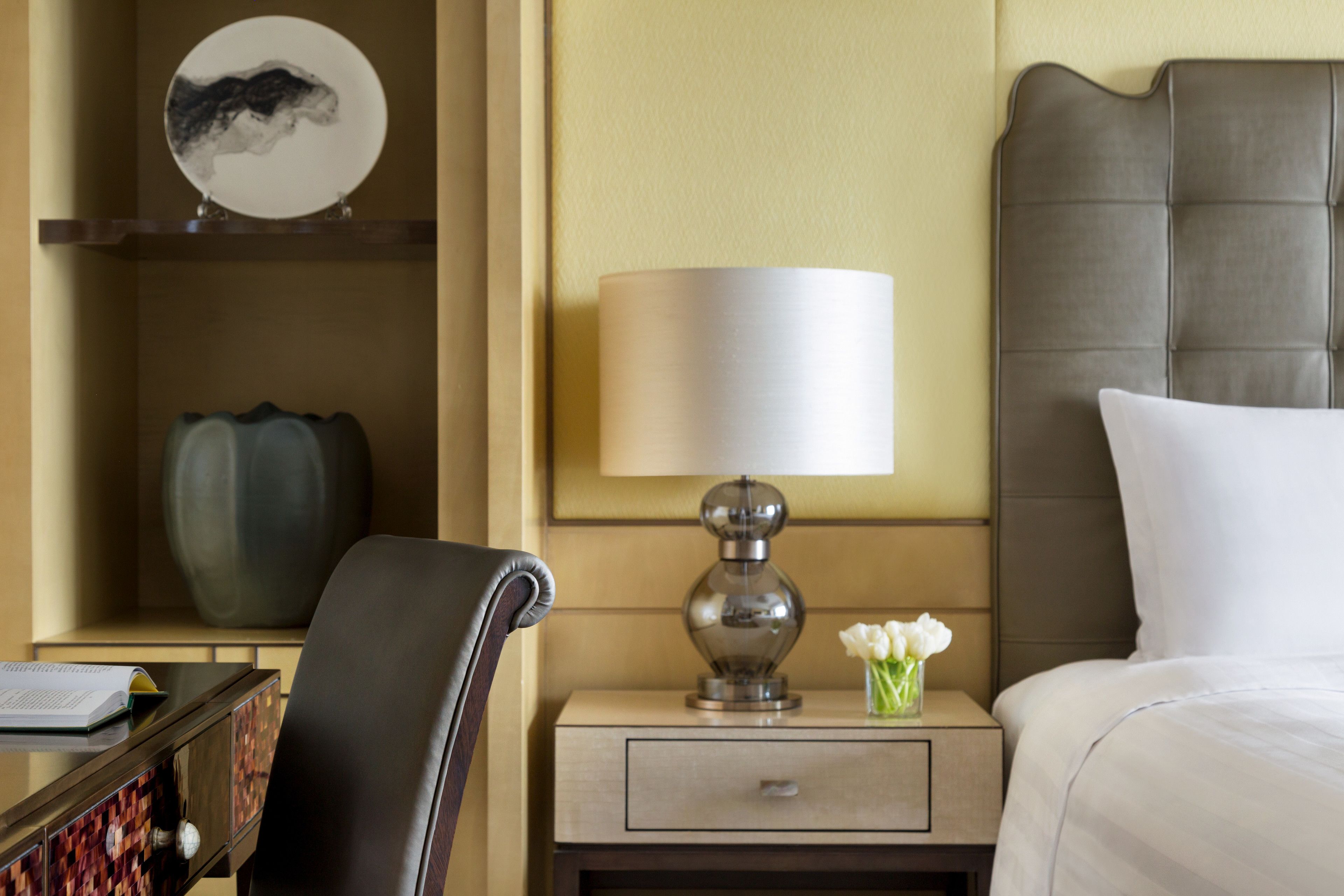 Una habitación de hotel con una cama, una mesita de noche con una lámpara, una silla, un escritorio con un libro y un plato decorativo en la pared.