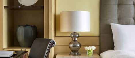 Una habitación de hotel con una cama, una mesita de noche con una lámpara, una silla, un escritorio con un libro y un plato decorativo en la pared.