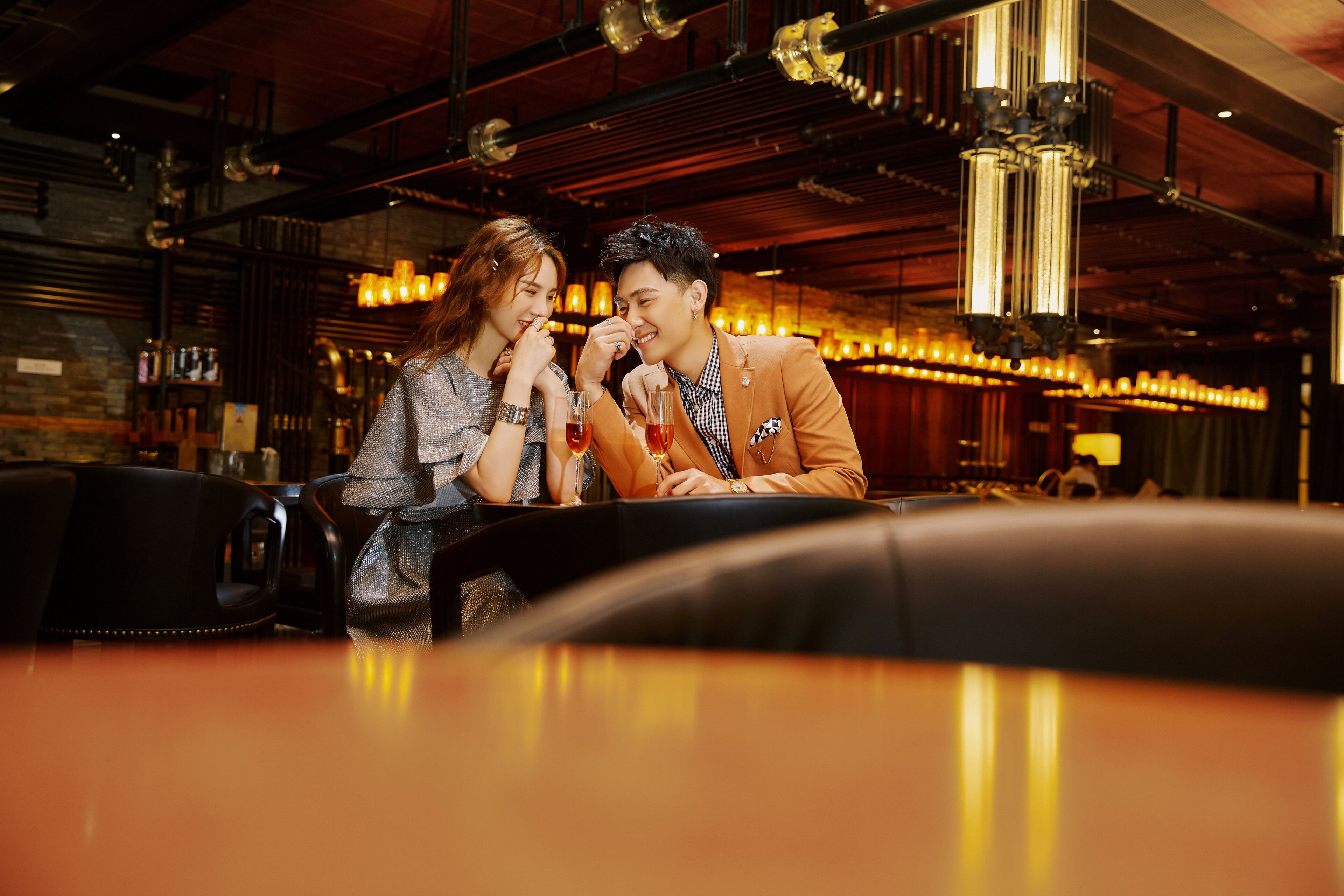 Un couple est assis au bar d’un établissement au décor industriel moderne, avec des tuyaux apparents et des luminaires suspendus.