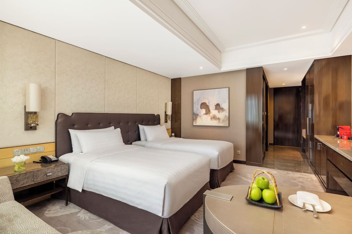 Ein Hotelzimmer mit einem großen Bett, Nachttischen, einem Schreibtisch, einem Sessel, einer Obstschale und einem Bild an der Wand.
