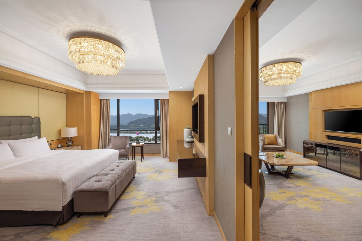 A hotel room with a large bed, a sofa, a TV, a dining table, and a view of mountains.