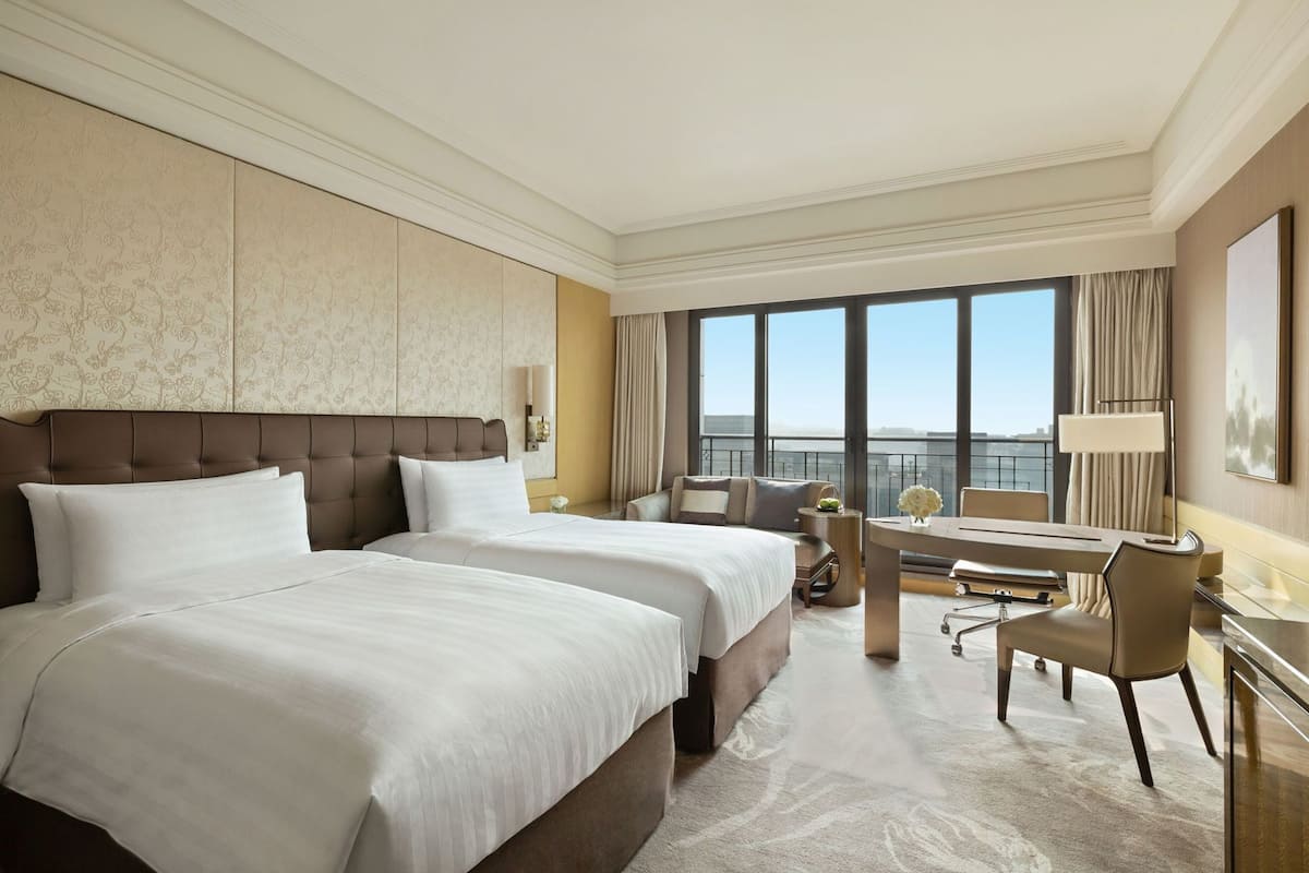 A hotel room with two beds, a desk, a chair, a large window with a view, and a decorative wall panel.