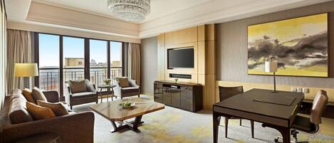 A modern hotel room with a sofa, armchairs, a dining table, a TV, and a large painting.