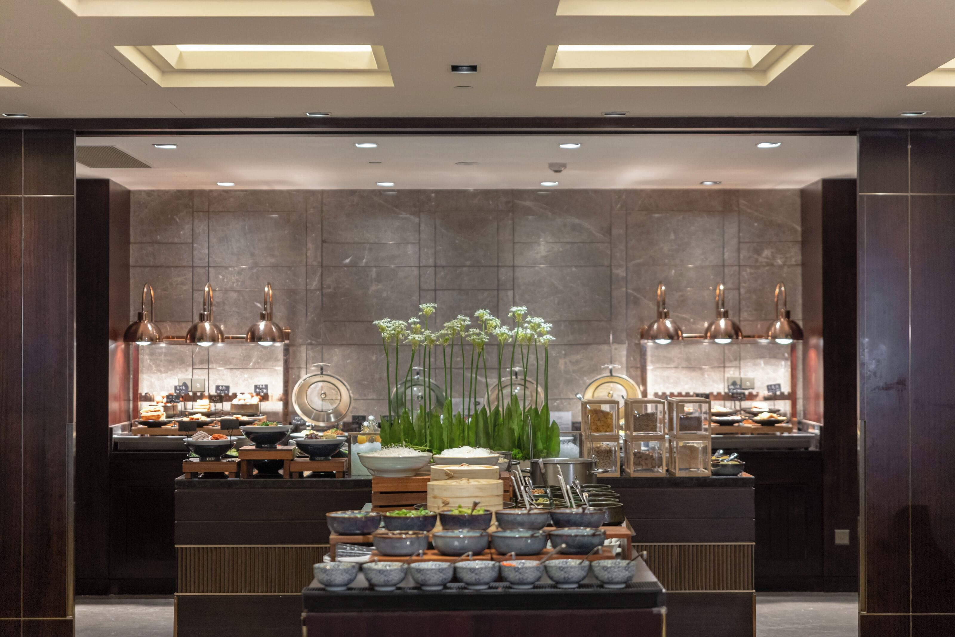 A buffet setup with various dishes and bowls, a central floral arrangement, and a modern interior with dark wood paneling and recessed lighting.