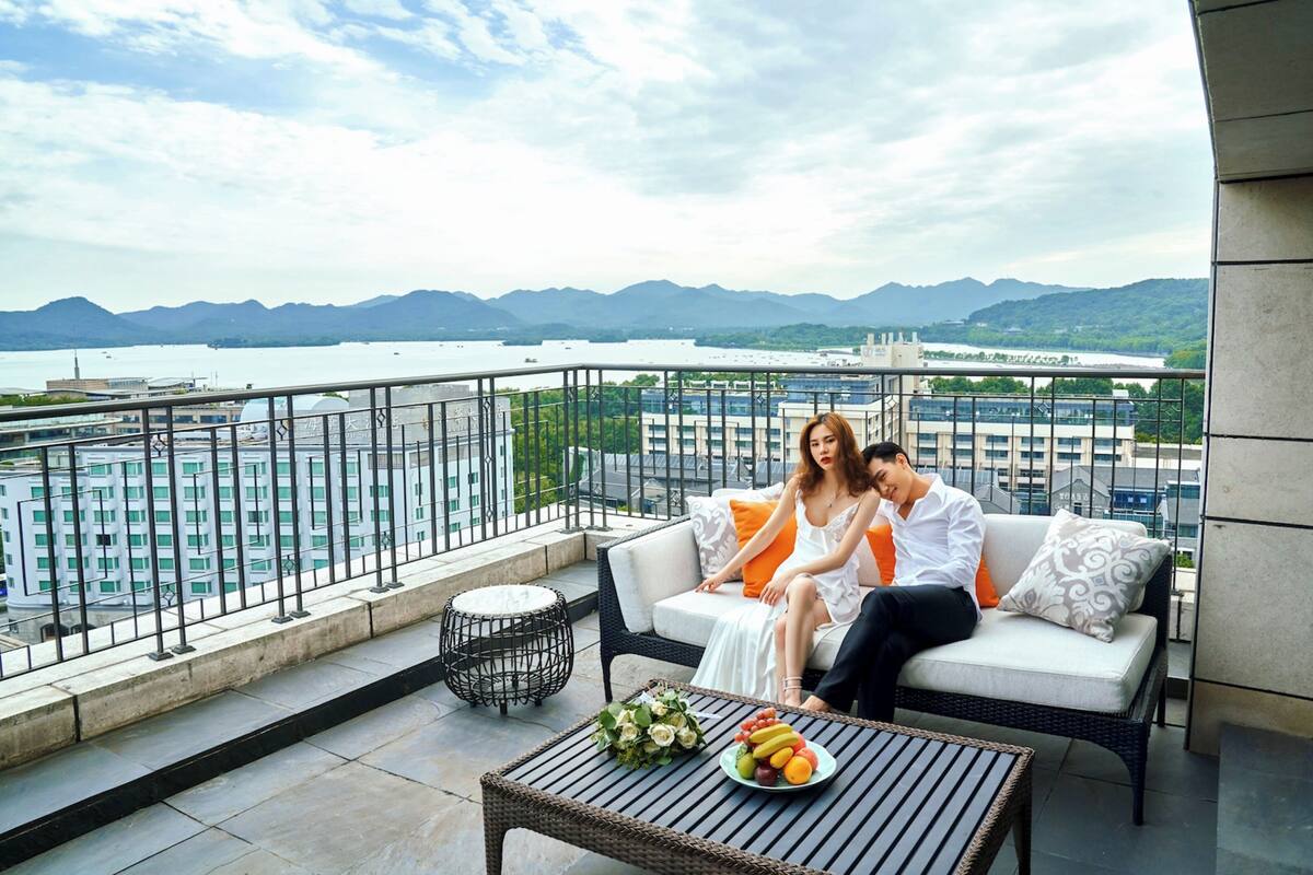 A balcony with a view of a body of water and mountains, a wicker sofa, a small round table with fruit, and a metal railing.