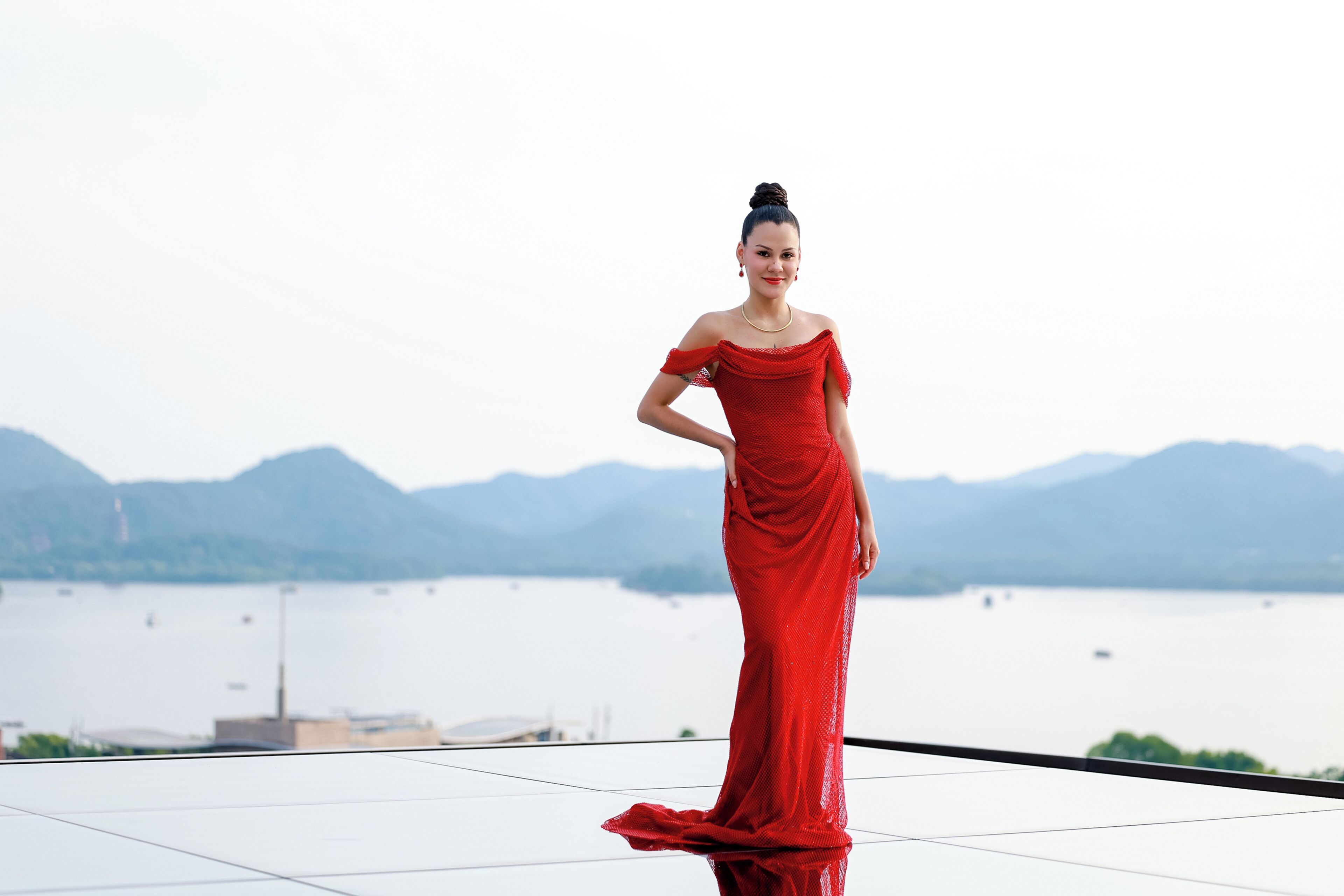 Eine Frau in einem roten Kleid steht auf einem Balkon mit einem See und Bergen im Hintergrund.