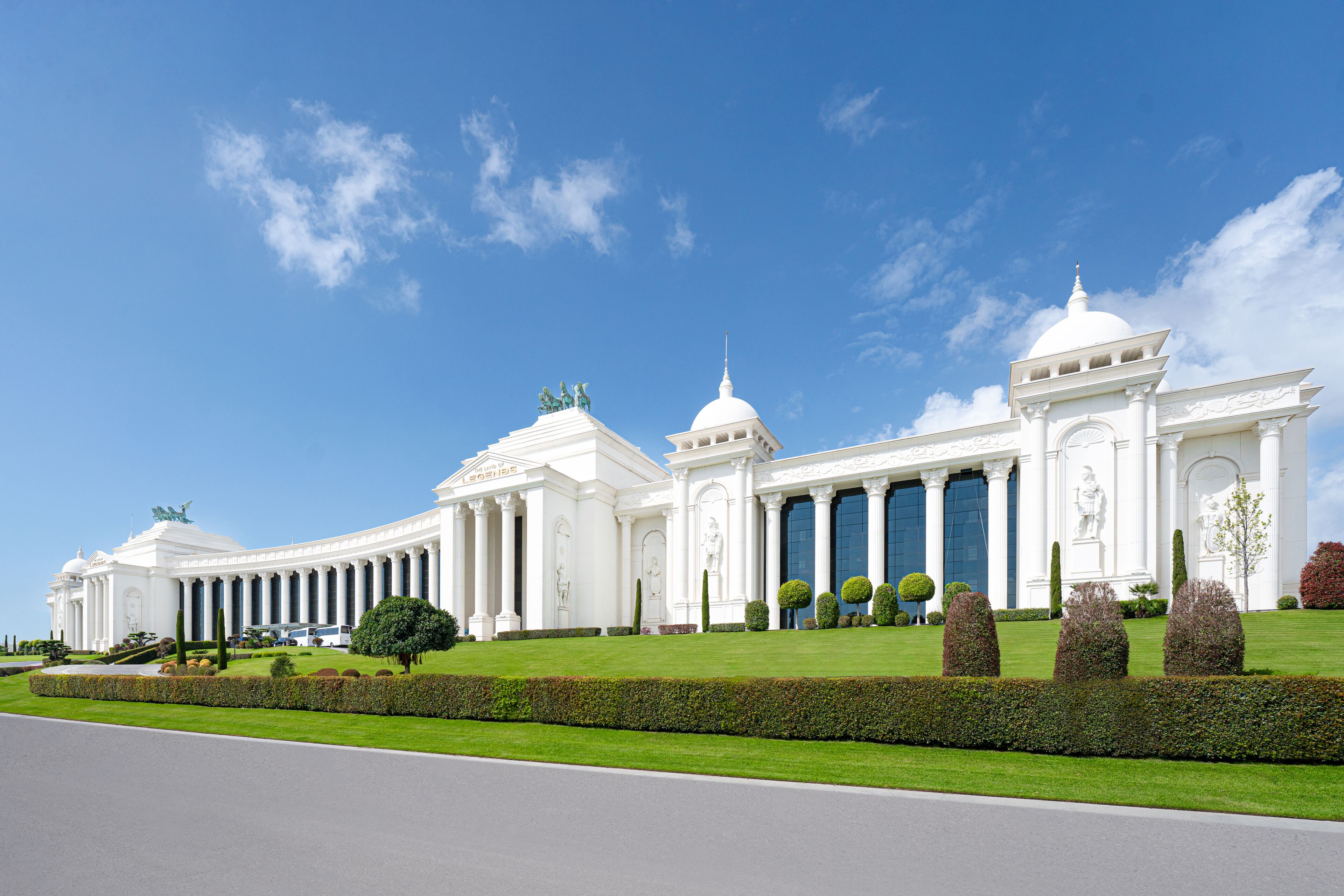 A large, white, classical-style building with columns and domes, surrounded by well-manicured lawns and hedges.