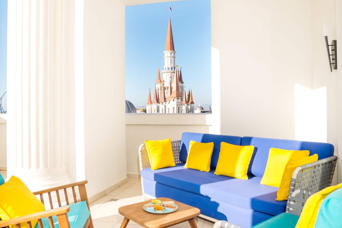 Ein helles Wohnzimmer mit einem blauen Sofa, gelben Kissen, einem hölzernen Schaukelstuhl und einem Korbstuhl. An der Wand hängt ein Gemälde eines Schlosses.