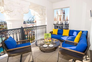 Ein Balkon mit Korbmöbeln, einem Glastisch und Blick auf ein schlossähnliches Gebäude.