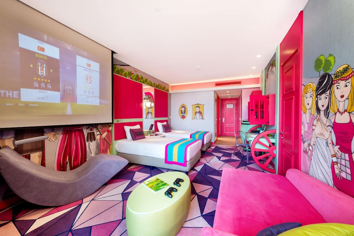Ein Hotelzimmer mit einem Bett, einer Couch, einem Fernseher, der Spielerstatistiken anzeigt, und einem farbenfrohen geometrischen Teppich.