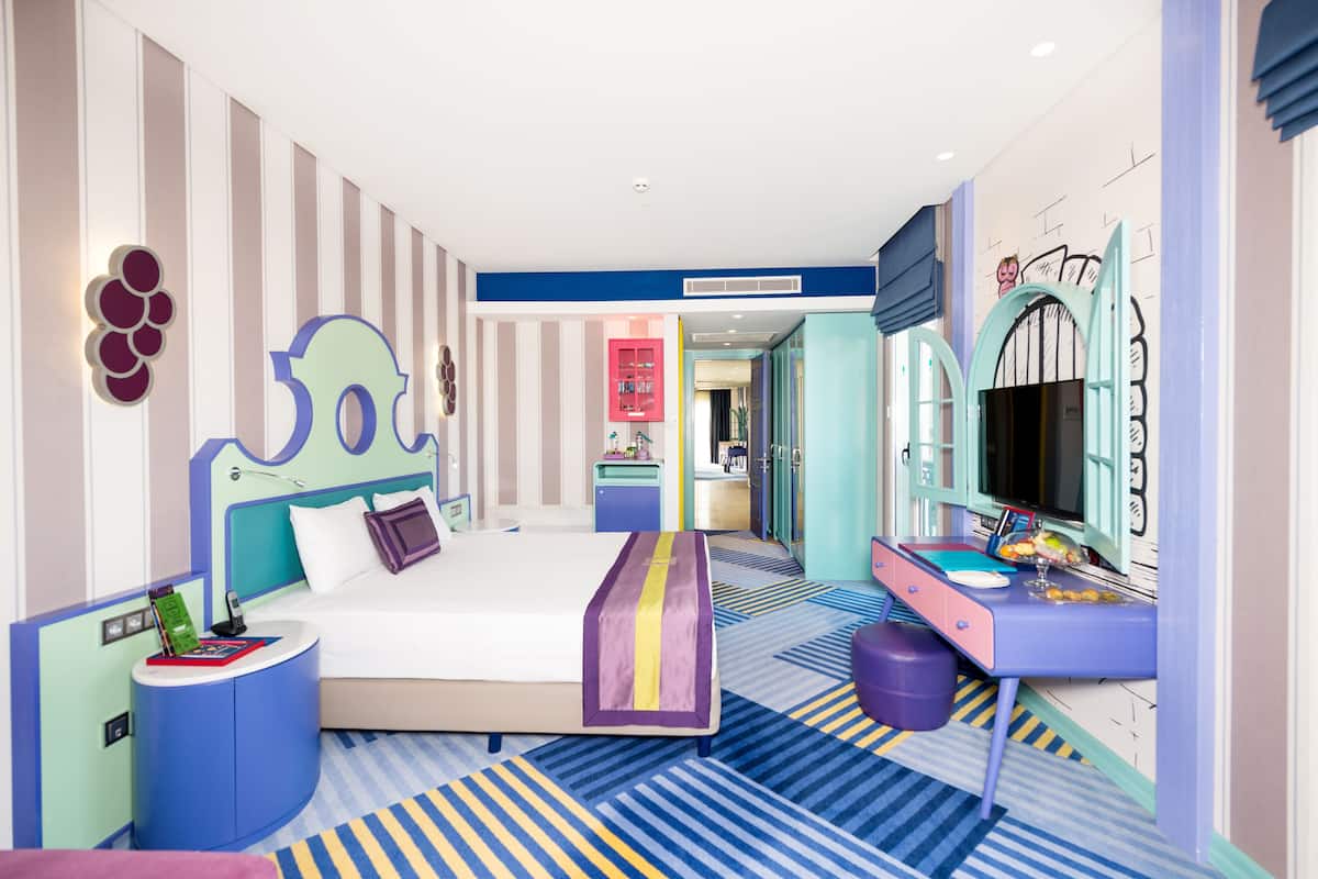 Ein modernes Hotelzimmer mit einem großen Bett, einem Schreibtisch, einem Fernseher und einem im Hintergrund sichtbaren Badezimmer.