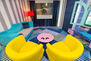 Ein Zimmer mit einer gestreiften Wand, einem blauen Sofa, einem runden pinkfarbenen Tisch mit zwei Spielcontrollern und einem Fernseher, der ein Rennspiel zeigt.