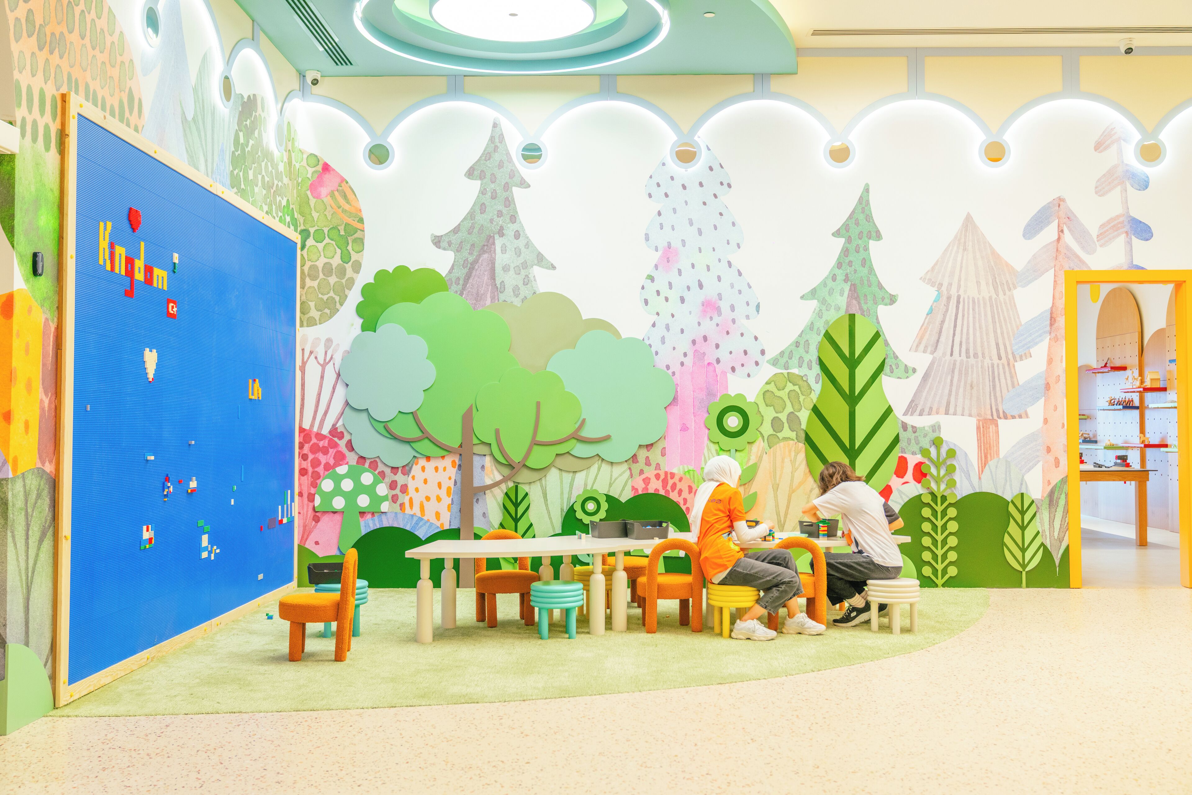 Un área de juegos infantiles con murales coloridos, una pared de juegos azul y una mesa con sillas.