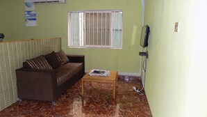 A living room with a brown sofa, a small wooden table, a window with metal bars, and a television mounted on the wall. - Posh Apartments Self Catering (Lagos)