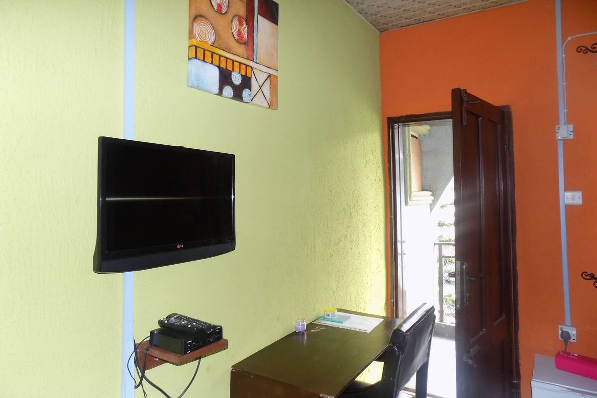 A room with a flat-screen TV mounted on the wall, a small shelf with a remote control, a table with a chair, and a door leading to another room.
