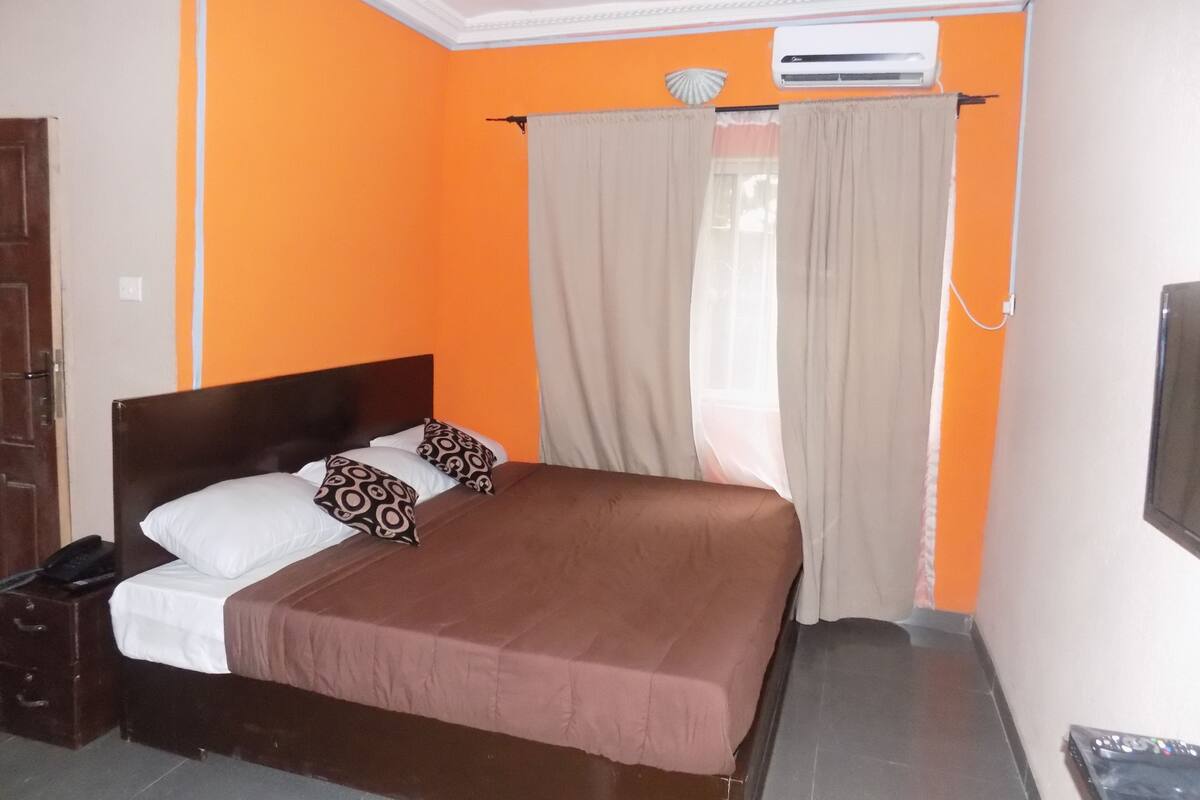 A bedroom with an orange wall, a brown bed, white and patterned pillows, a brown bedspread, a window with beige curtains, and an air conditioning unit.