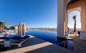 Une piscine à débordement avec des chaises longues et des parasols, offrant une vue sur une plage et la mer.