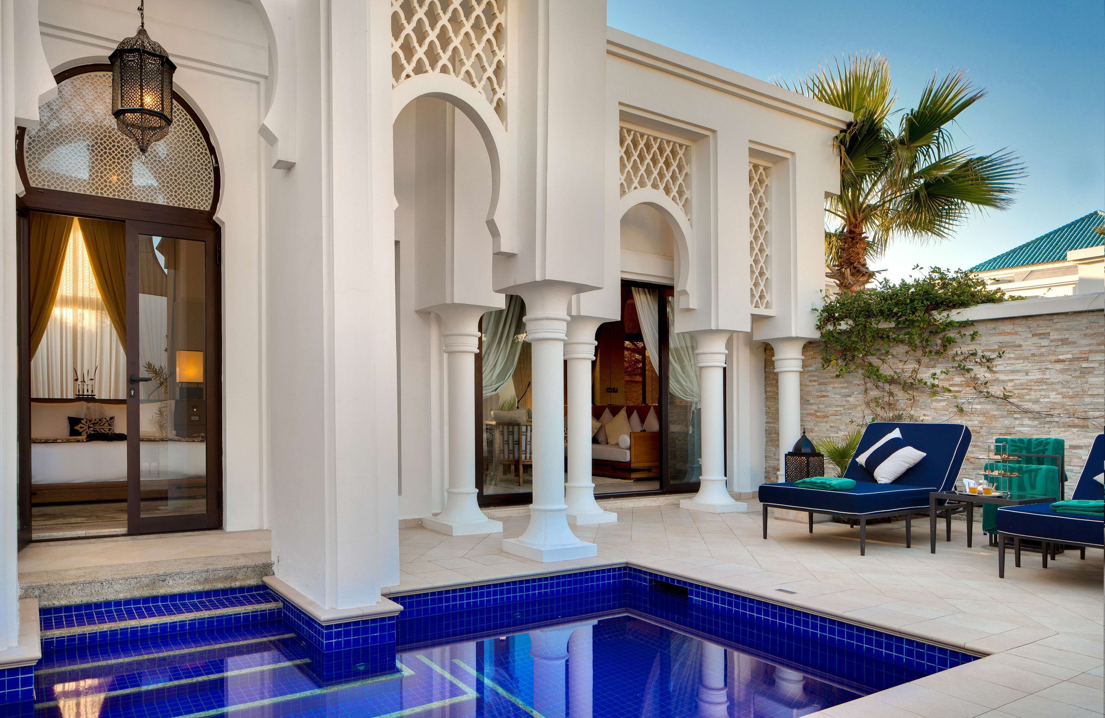 Un espace aménagé au bord de la piscine, avec une piscine aux carreaux bleus, des colonnes blanches et un coin salon équipé de chaises longues.