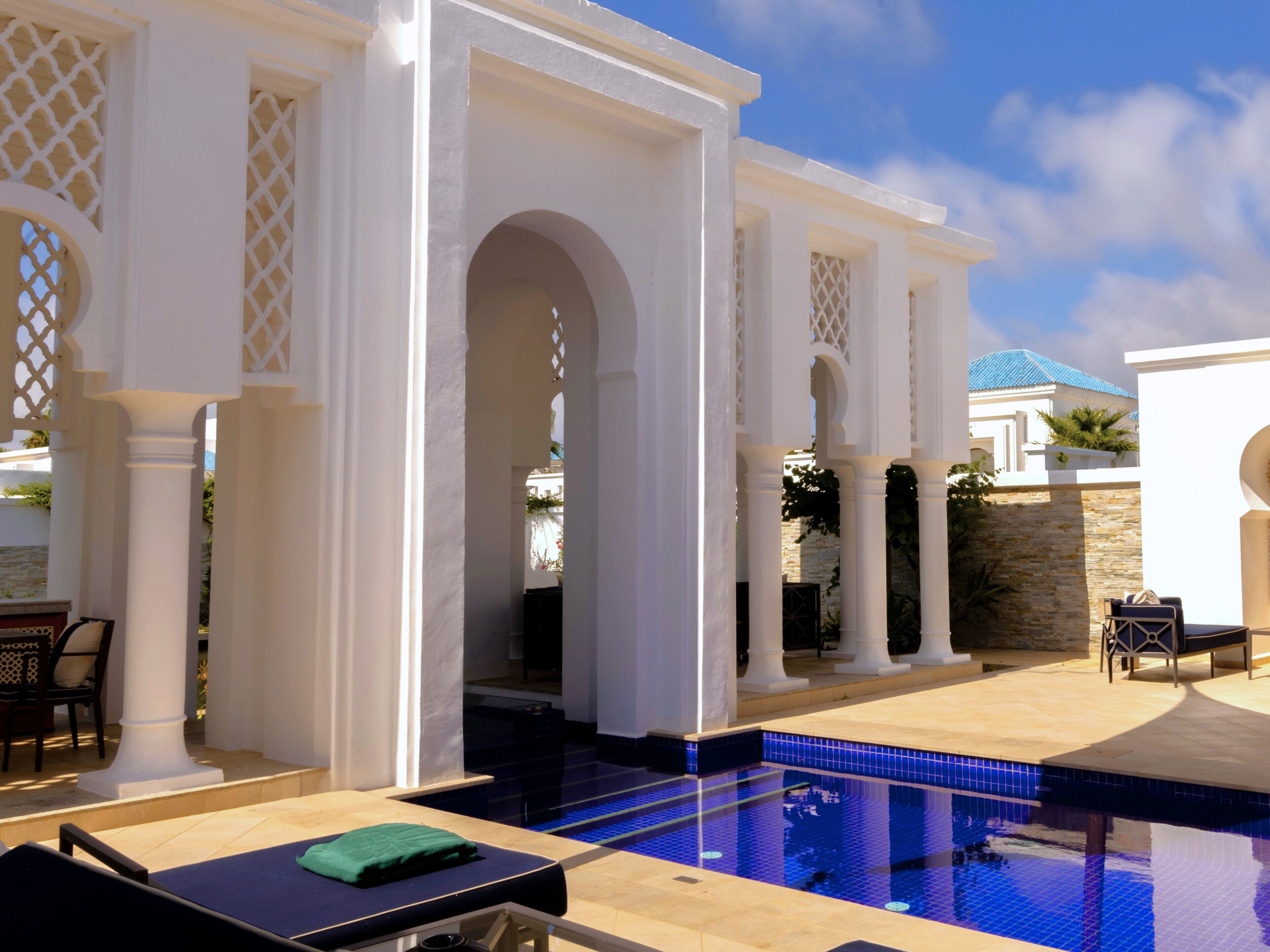 Un área de piscina con una piscina azul cristalina, columnas blancas y un muro de piedra.