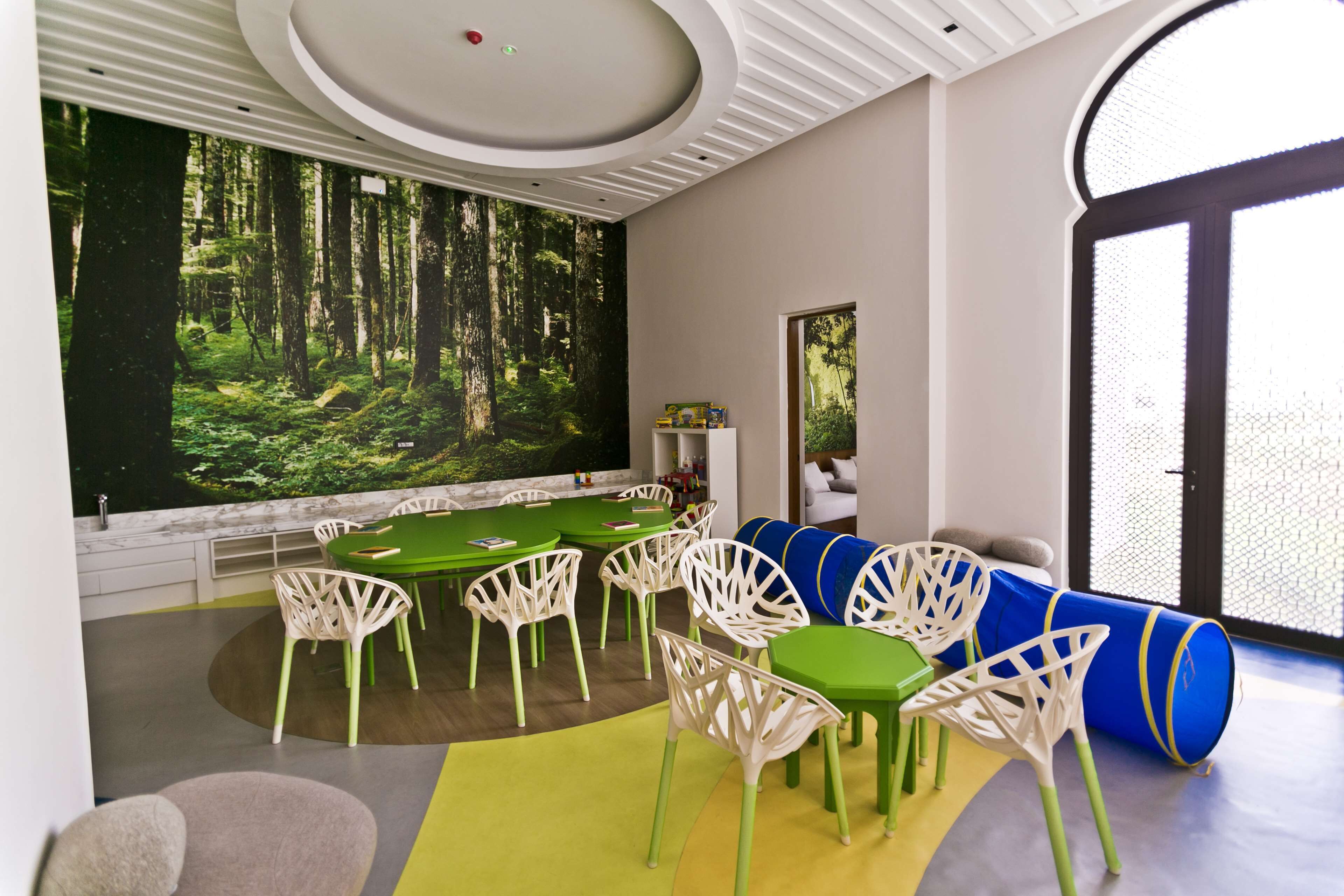 Une pièce moderne avec une table verte, des chaises blanches et une fresque représentant une forêt sur le mur.