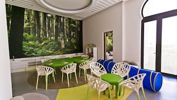 Une pièce moderne avec une table verte, des chaises blanches et une fresque représentant une forêt sur le mur.