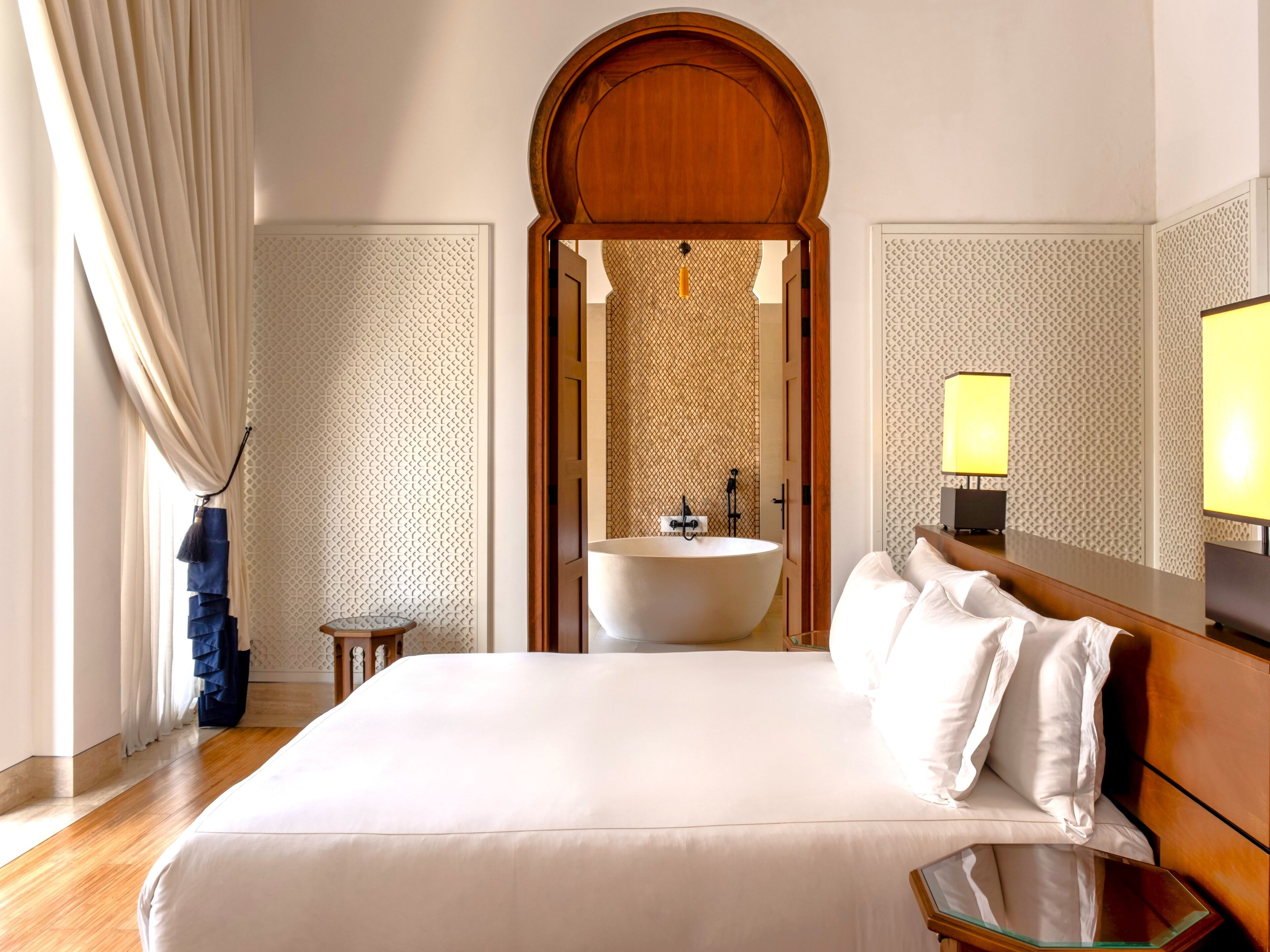 Ein Hotelzimmer mit einem großen Bett, einem hölzernen Kopfteil, einem runden Spiegel, einem Nachttisch, einem kleinen runden Tisch, einem großen runden Spiegel, einem Waschbecken und einem Fenster mit Vorhängen.