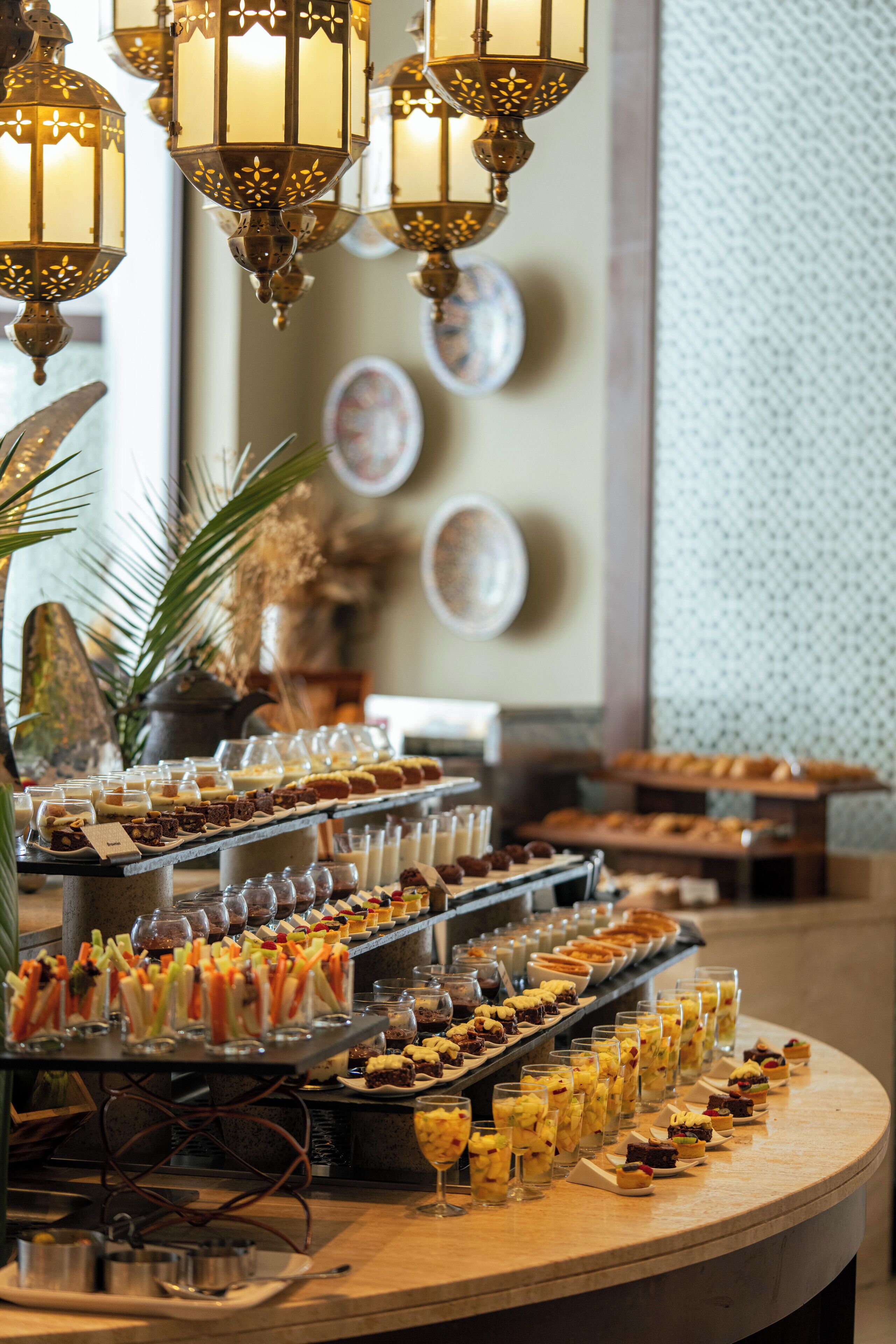 Un bufé con diversos postres y bebidas, una pared decorativa con platos y faroles colgantes.