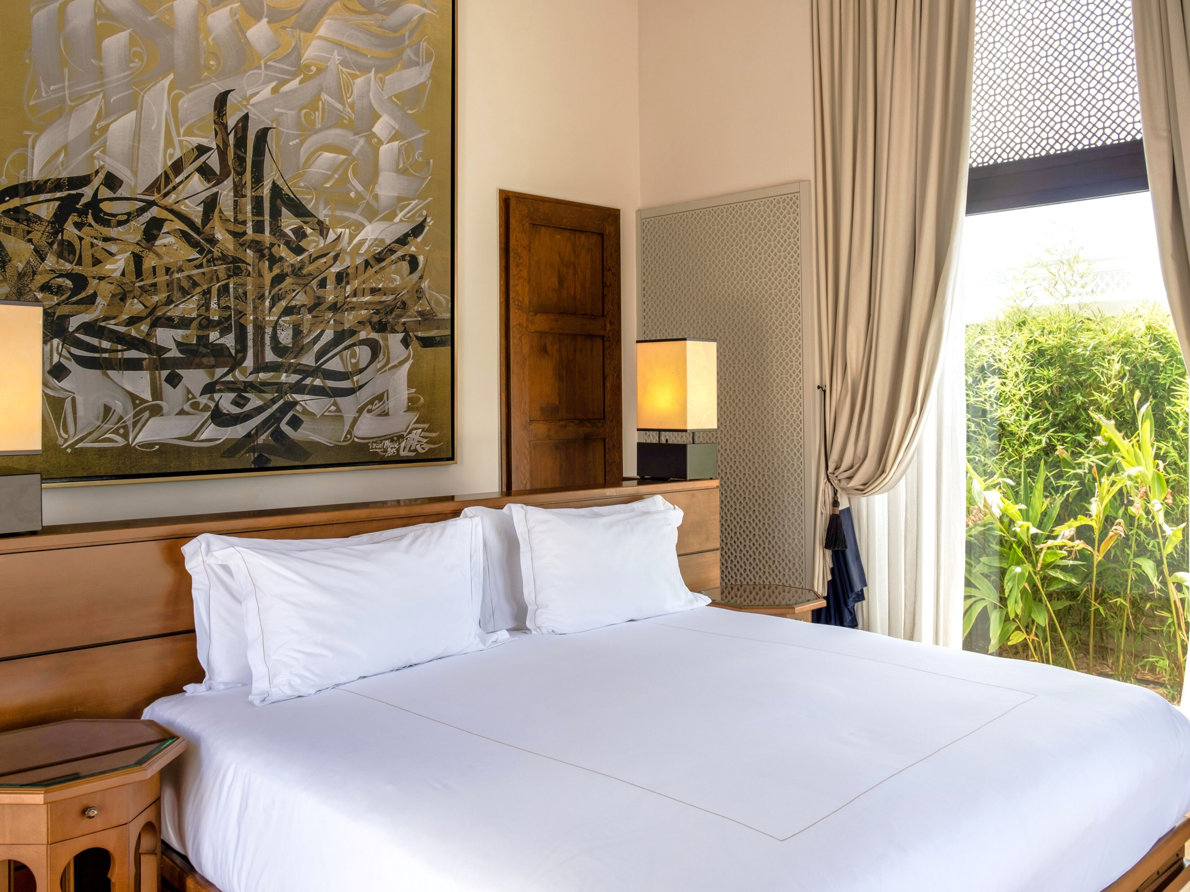 A hotel room with a large bed, two bedside lamps, a wooden headboard, a large abstract painting, a wooden wardrobe, and a window with curtains.