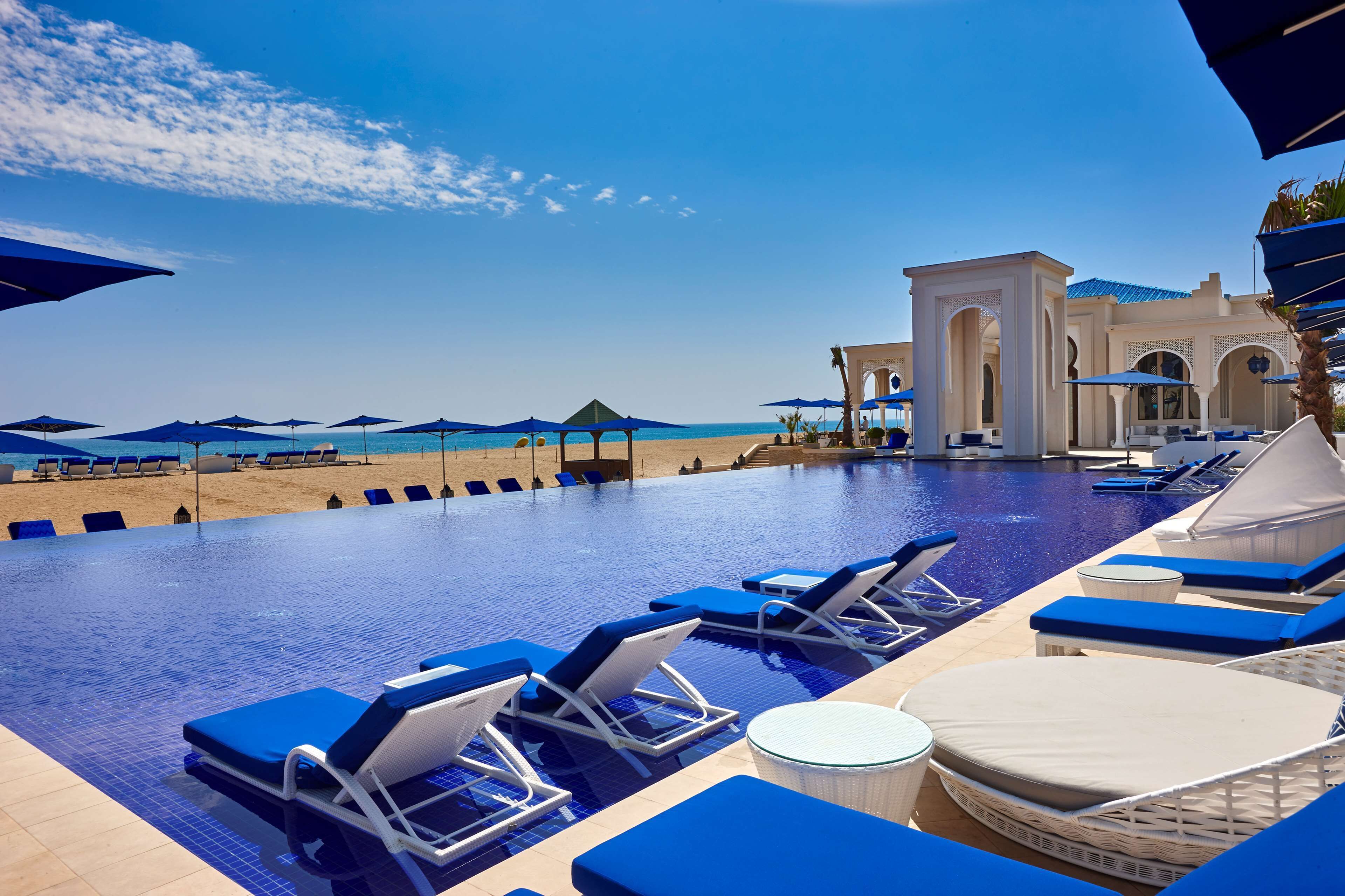 Un área junto a la piscina con sillas de descanso y sombrillas, vista despejada de la playa y un edificio con arcos al fondo.