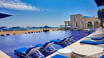Un área junto a la piscina con sillas de descanso y sombrillas, vista despejada de la playa y un edificio con arcos al fondo.