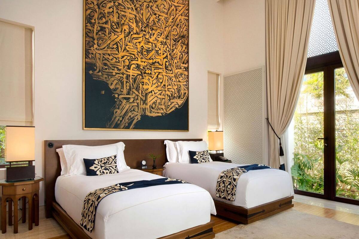 Ein Hotelzimmer mit zwei Betten, einem großen Kunstwerk an der Wand, Blick auf Grünfläche draußen und einem Fenster mit Vorhängen.