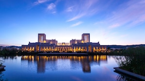 Un edificio grande e iluminado con una fachada simétrica y dos torres destacadas, reflejado en una masa de agua tranquila.