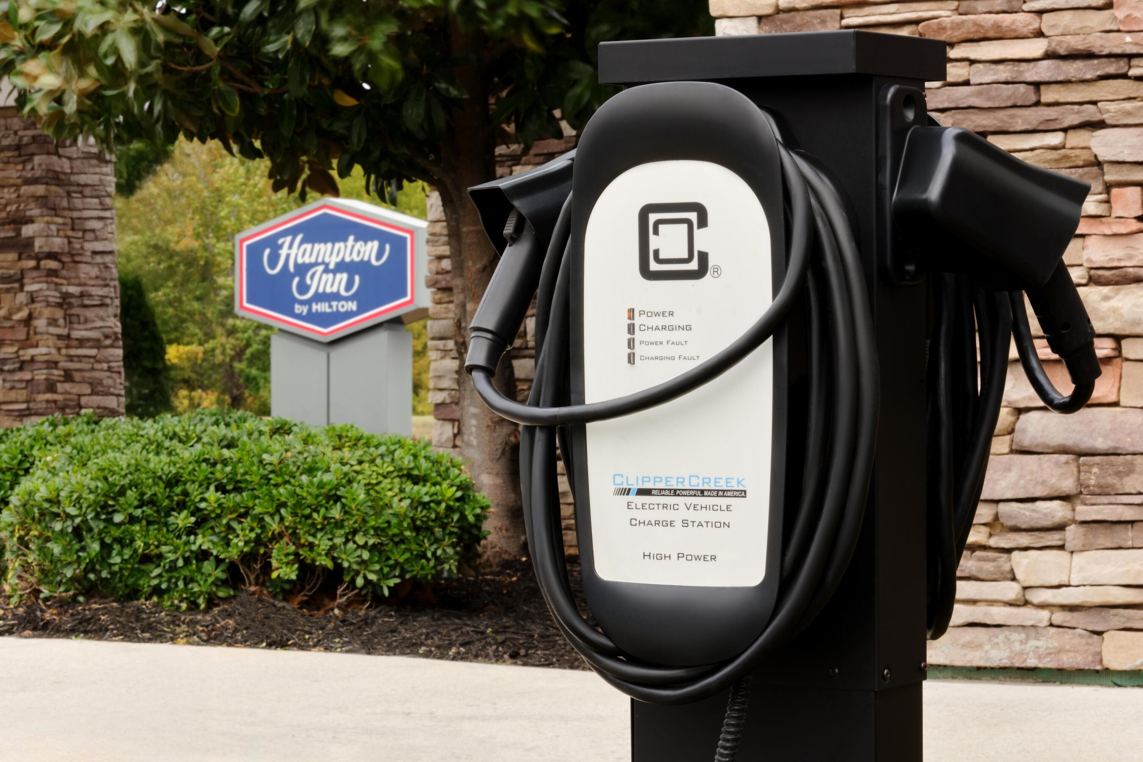 A Hampton Inn by Hilton electric vehicle charging station with a stone wall and greenery in the background.