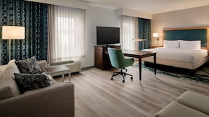 A hotel room with a bed, a desk with a flat-screen TV, a green armchair, a sofa, and a lamp.