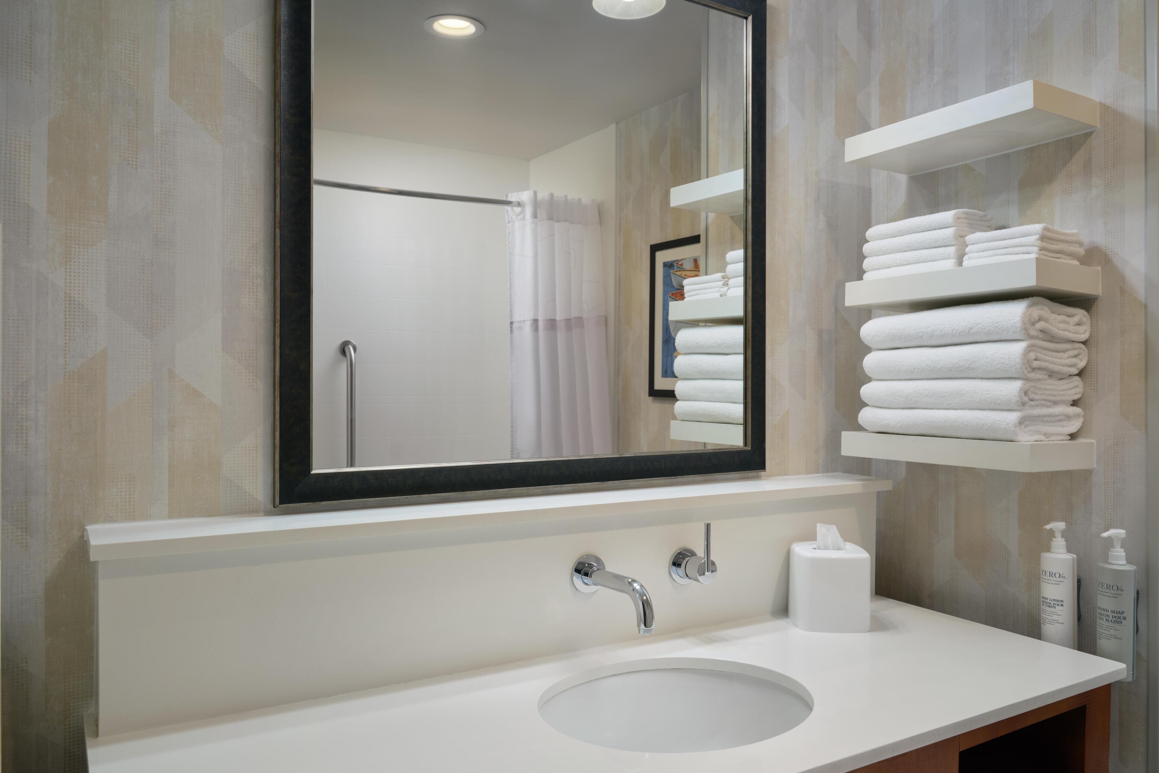 Une salle de bain moderne avec un grand miroir, un lavabo blanc et des serviettes soigneusement pliées et rangées sur des étagères.