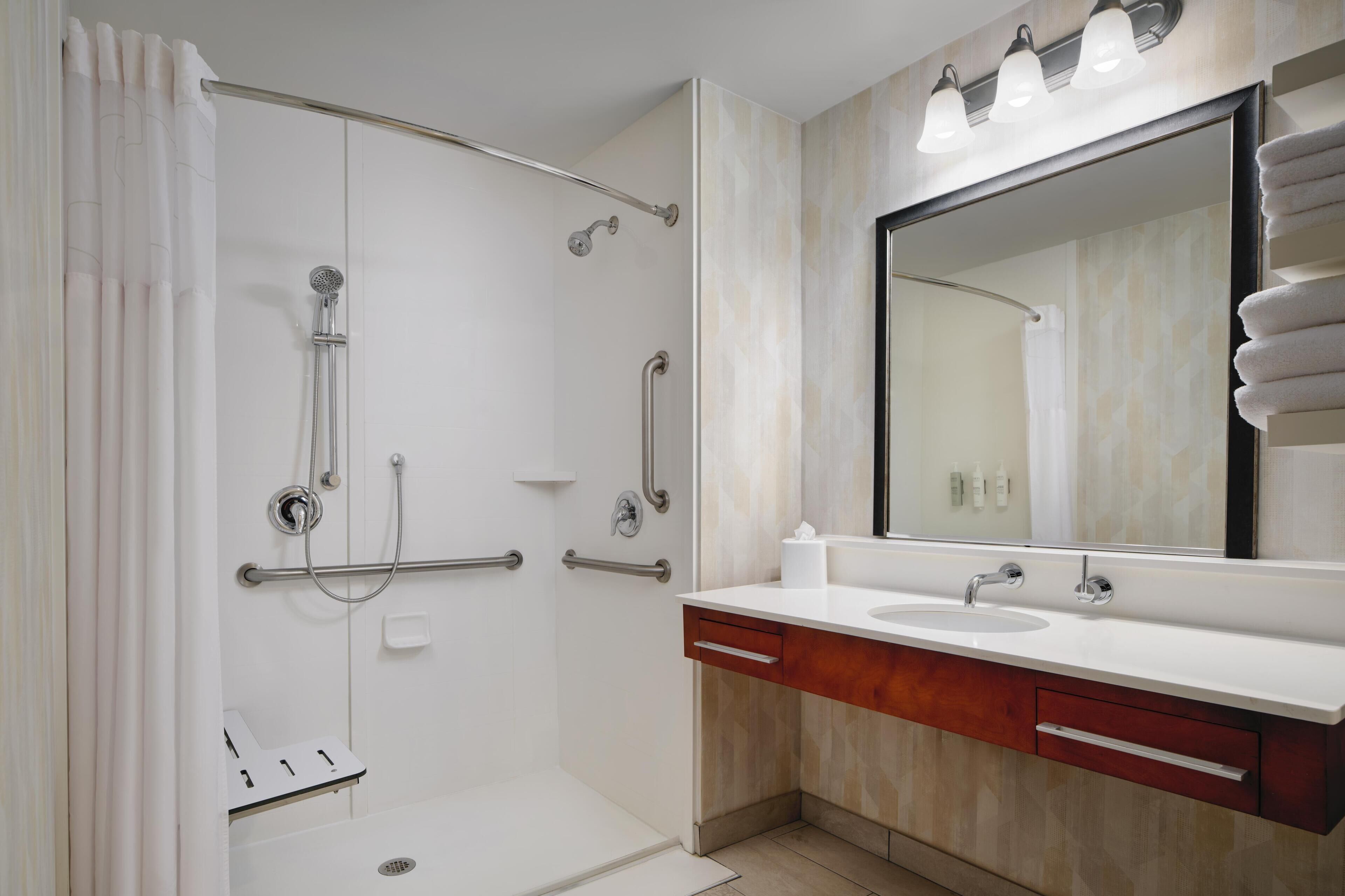 Un baño moderno con ducha a nivel, barras de apoyo, un espejo grande y un tocador con lavamanos y perchero.