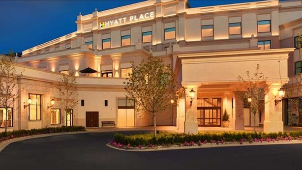 A large, well-lit hotel with a prominent 'Hyatt Place' sign, featuring a symmetrical facade, multiple windows, and a landscaped entrance area.