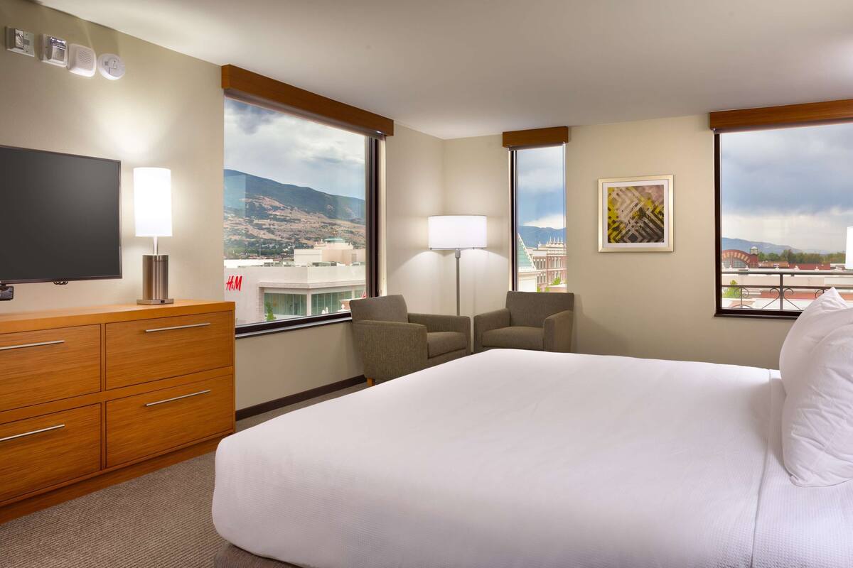 Une chambre d’hôtel avec un grand lit, une télévision, un bureau et une chaise. Les fenêtres offrent une vue sur les bâtiments et les montagnes.