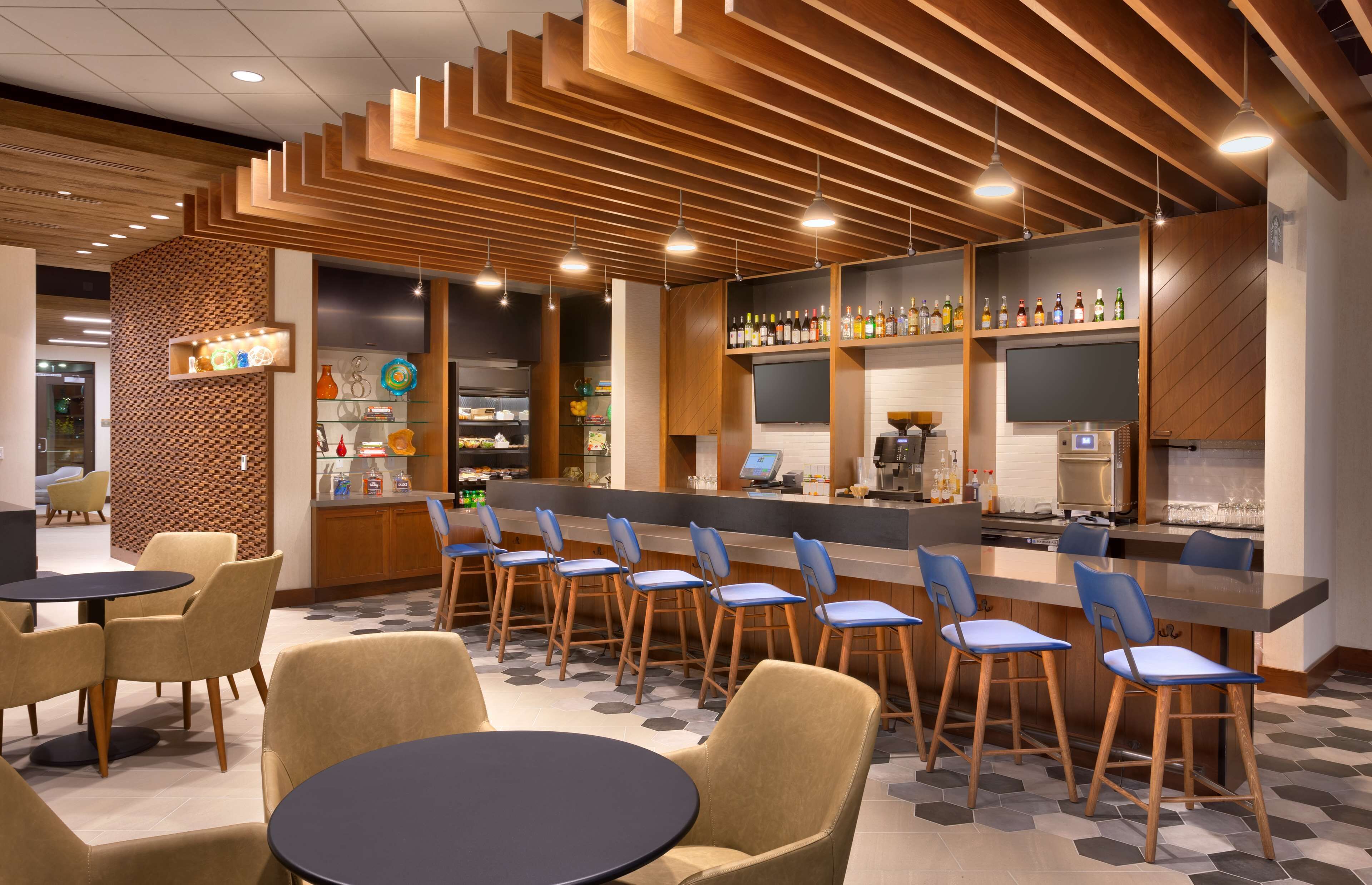 Un área de bar moderna con una barra larga, distintas opciones de asientos, incluyendo taburetes y mesas redondas, y una variedad de botellas en estantes.