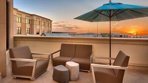 A modern outdoor seating area with a sofa, armchairs, and ottomans, featuring a large umbrella and a view of a building with a glass facade.